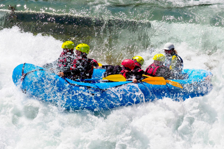 Group wearing helmets and life vests white-water rafting in blue inflatable boat.